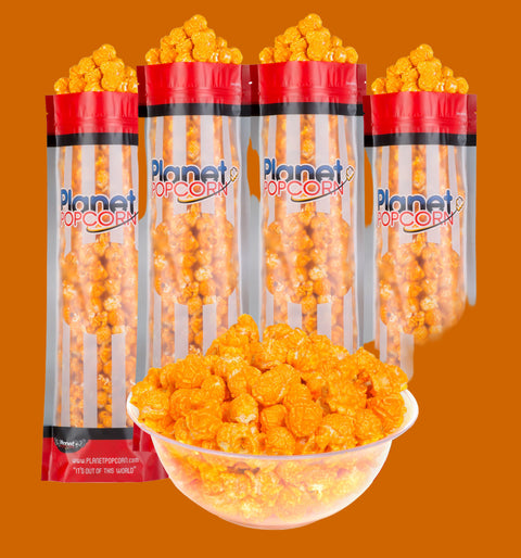 Four bags of Planet Popcorn's Spicy Cheddar Big Dipper popcorn, with one large clear bowl filled with popcorn in the foreground.