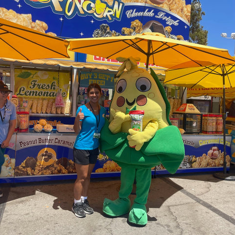 Person in a green mascot costume standing next to a lemon-themed stand at a the Orange County Fair
